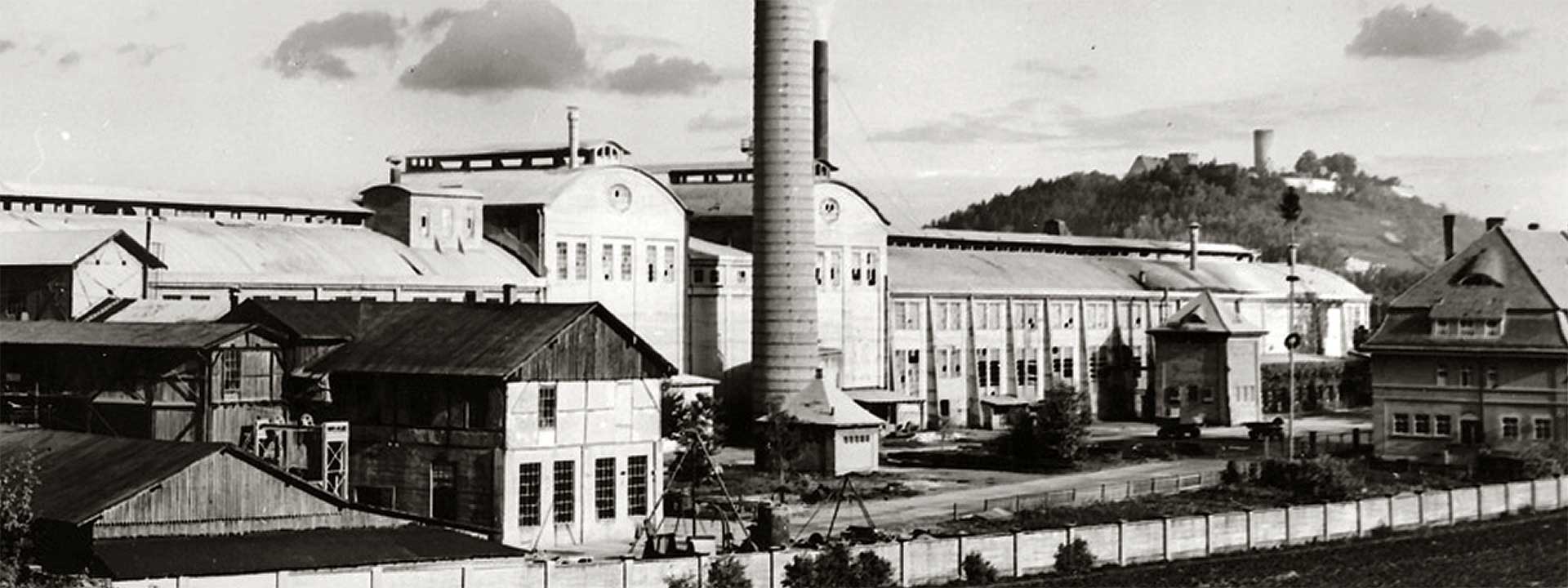 Historisches Schwarz-Weiß-Foto eines Industriekomplexes mit markantem Schornstein vor Hügelkulisse