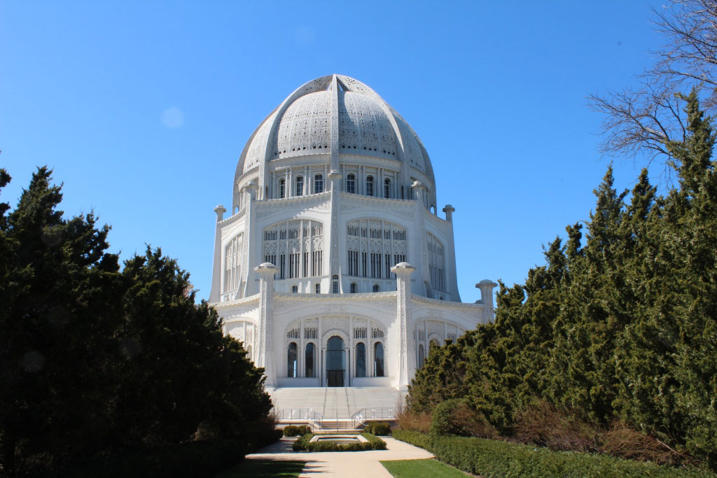 A large, white building with a distinctive, tall dome structure at the center, surrounded by clear blue skies. The architecture of the building is notable for its grandeur and symmetry. Trees line either side of a central pathway leading up to the building, suggesting a formal garden setting.