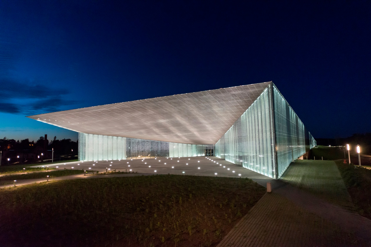 A modern, white building with a distinctive, sloping roof and a large glass facade. The building is illuminated at night and surrounded by a paved plaza.