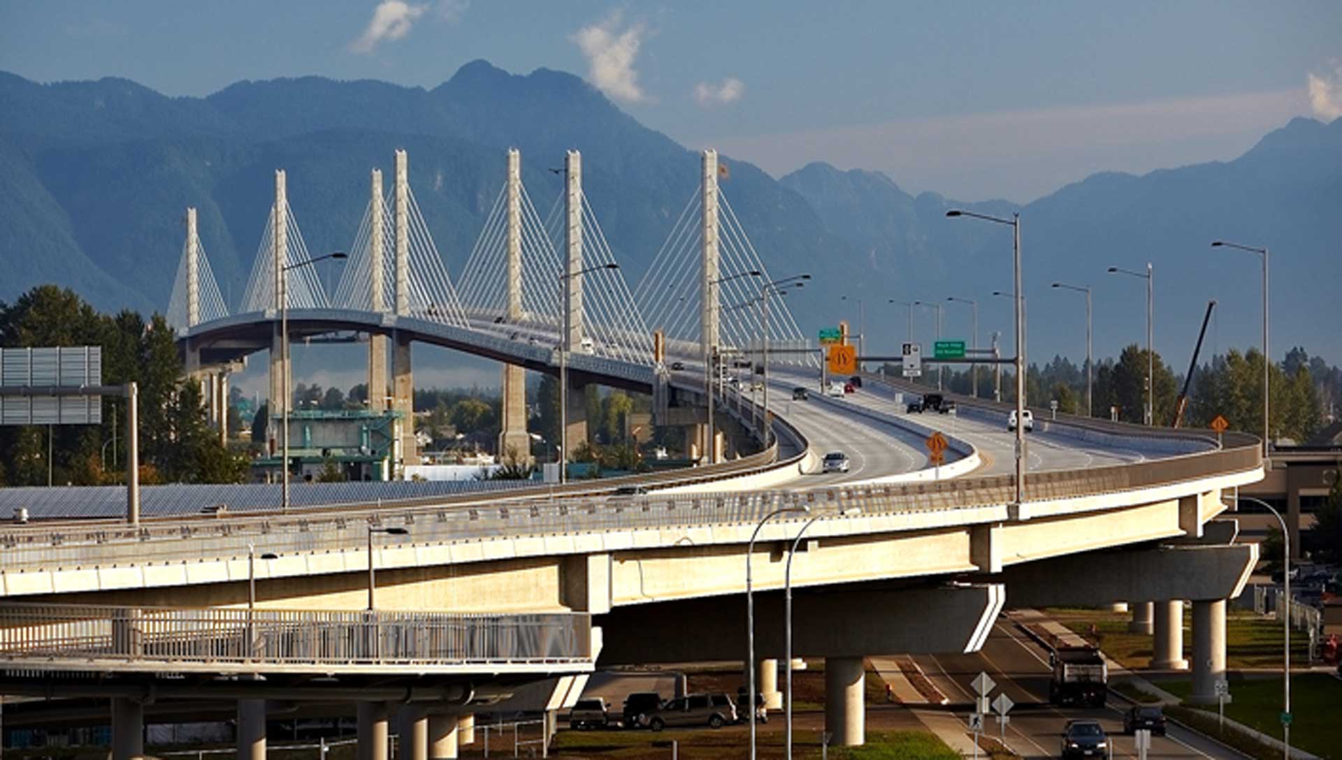 A modern bridge with multiple lanes, featuring distinctive white supports that rise above the roadway in a series of tall, thin spires.