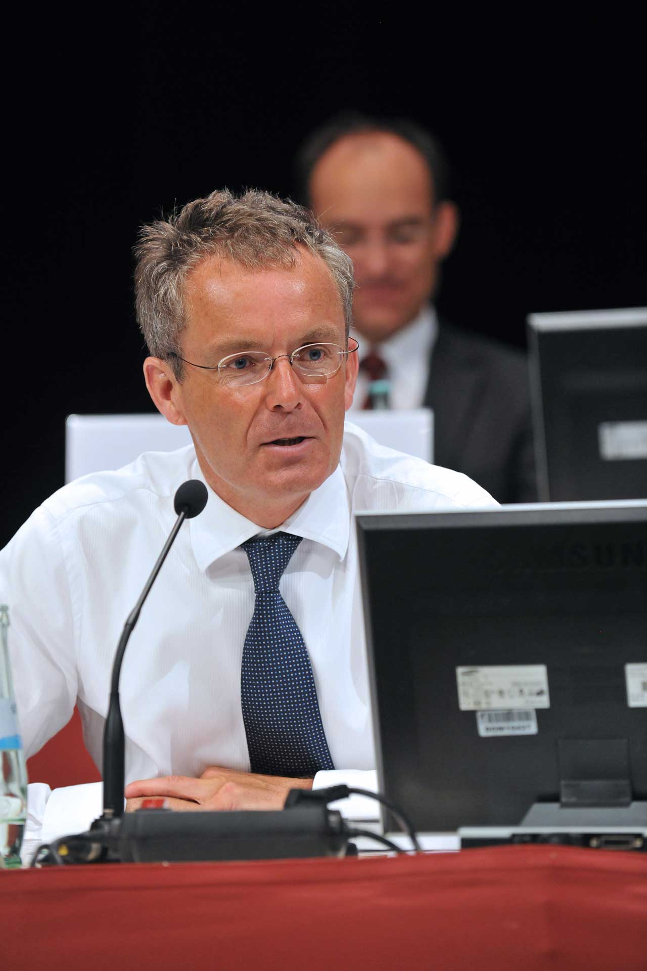 grey-haired man in a white shirt and tie behind a microphone and monitor