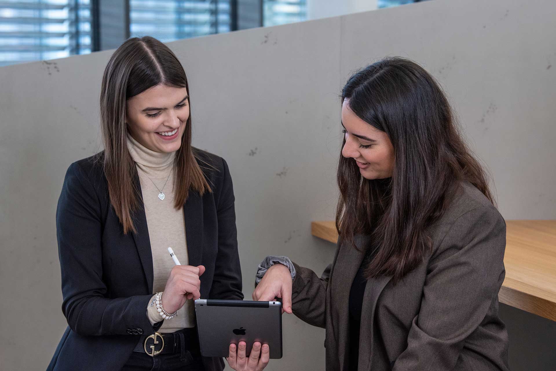 Zwei Frauen in Businesskleidung unterhalten sich und schauen dabei in ein Tablet, das die linke davon in der Hand hält