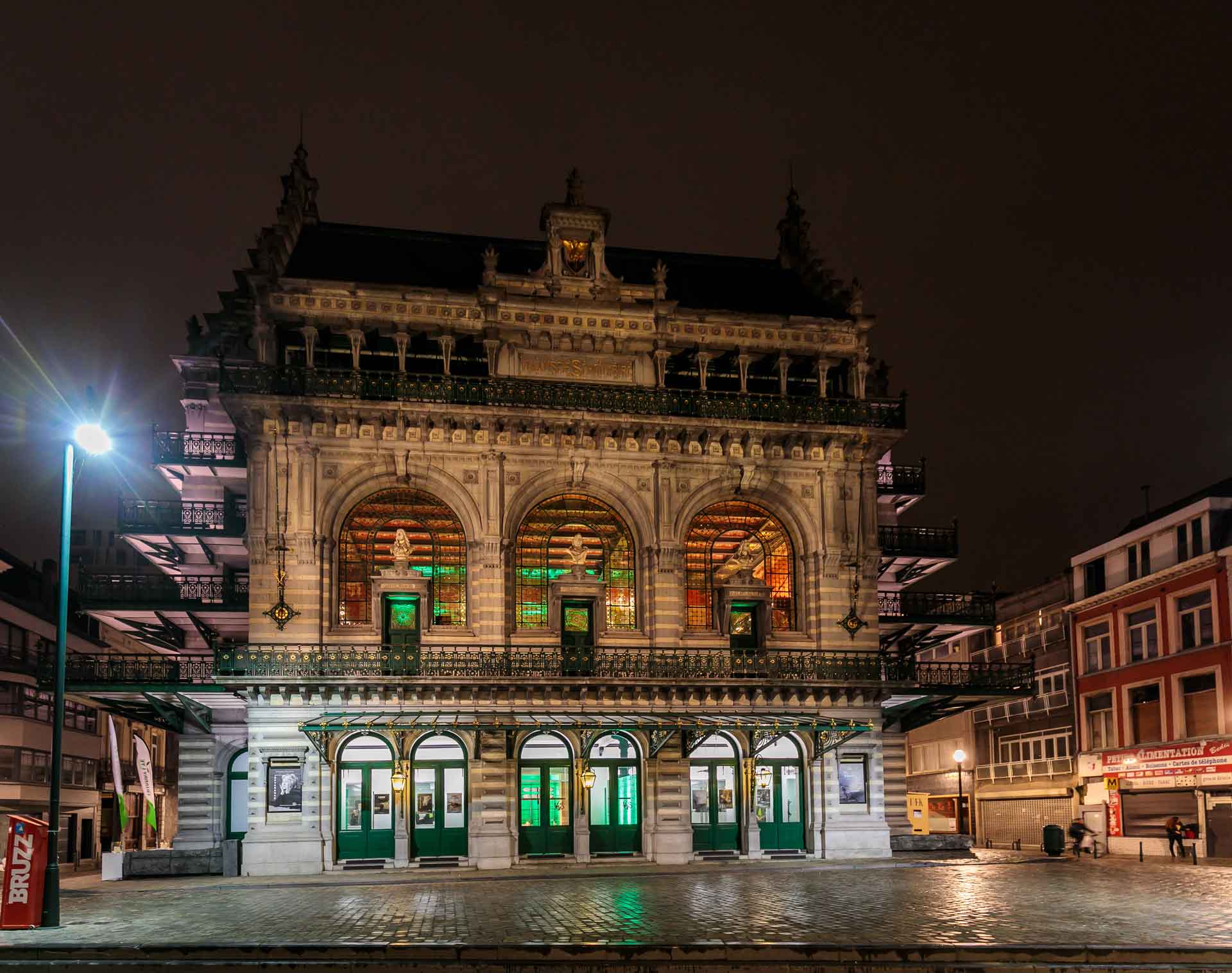 A grand, illuminated theater building with arched windows and ornate detailing. The building is lit up at night, creating a striking contrast against the dark sky.