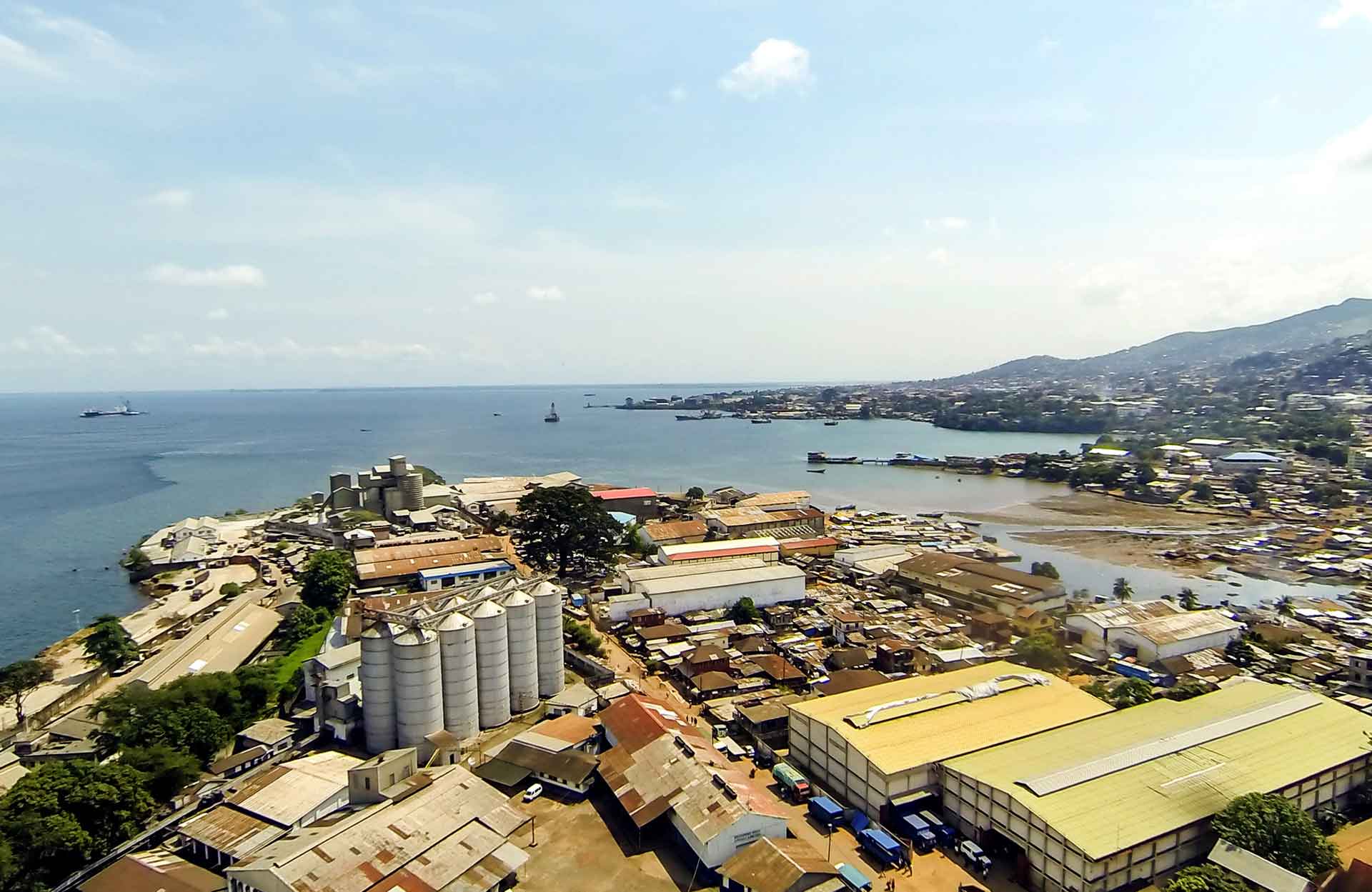 An aerial view of a coastal city with a port and industrial area. There are several buildings, including a silo, and a large body of water with ships in the background.