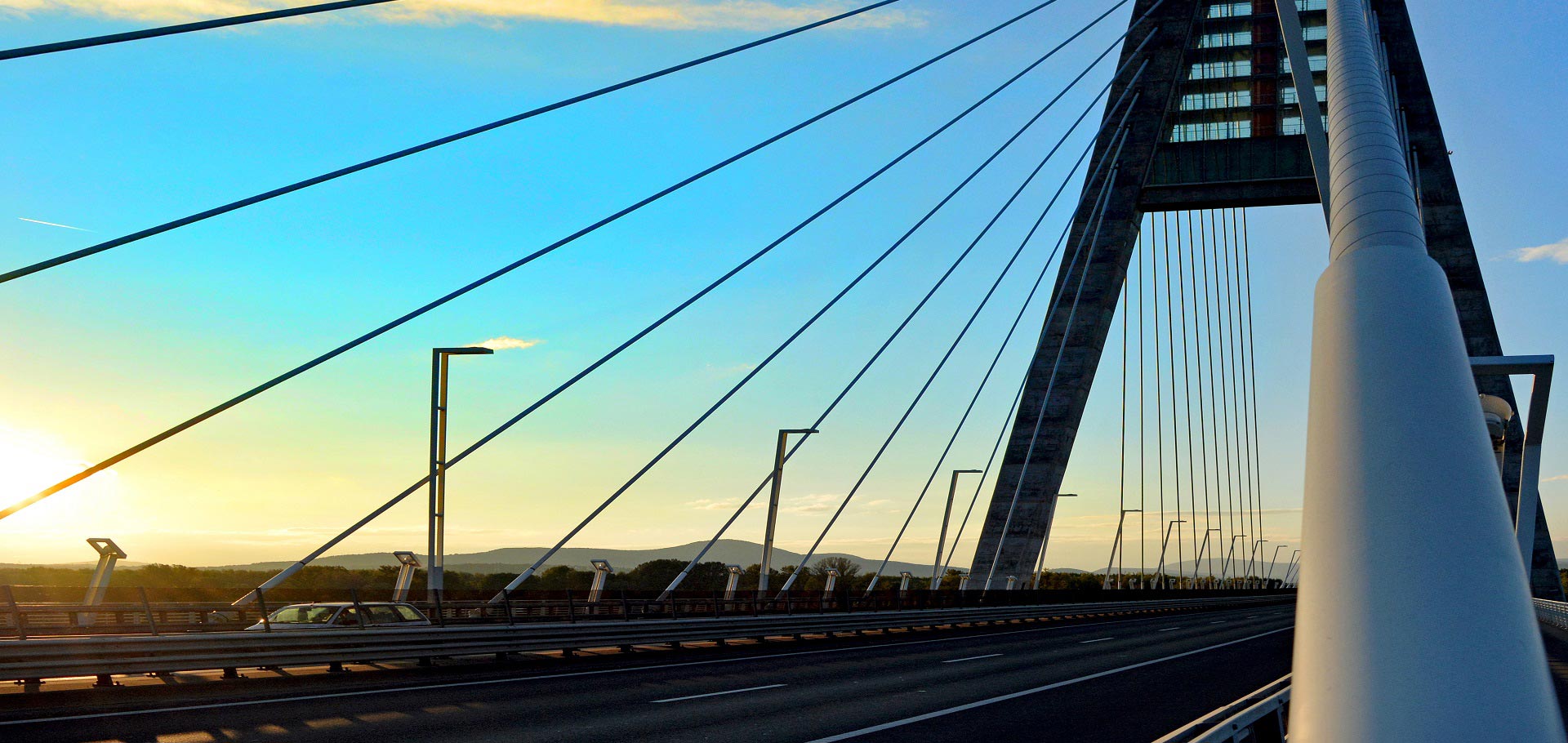 Eine moderne Schrägseilbrücke mit einem weißen Turm und zahlreichen Seilen, die sich von ihm zur Fahrbahn erstrecken. Die Sonne geht im Hintergrund unter und wirft ein warmes Licht auf die Brücke.