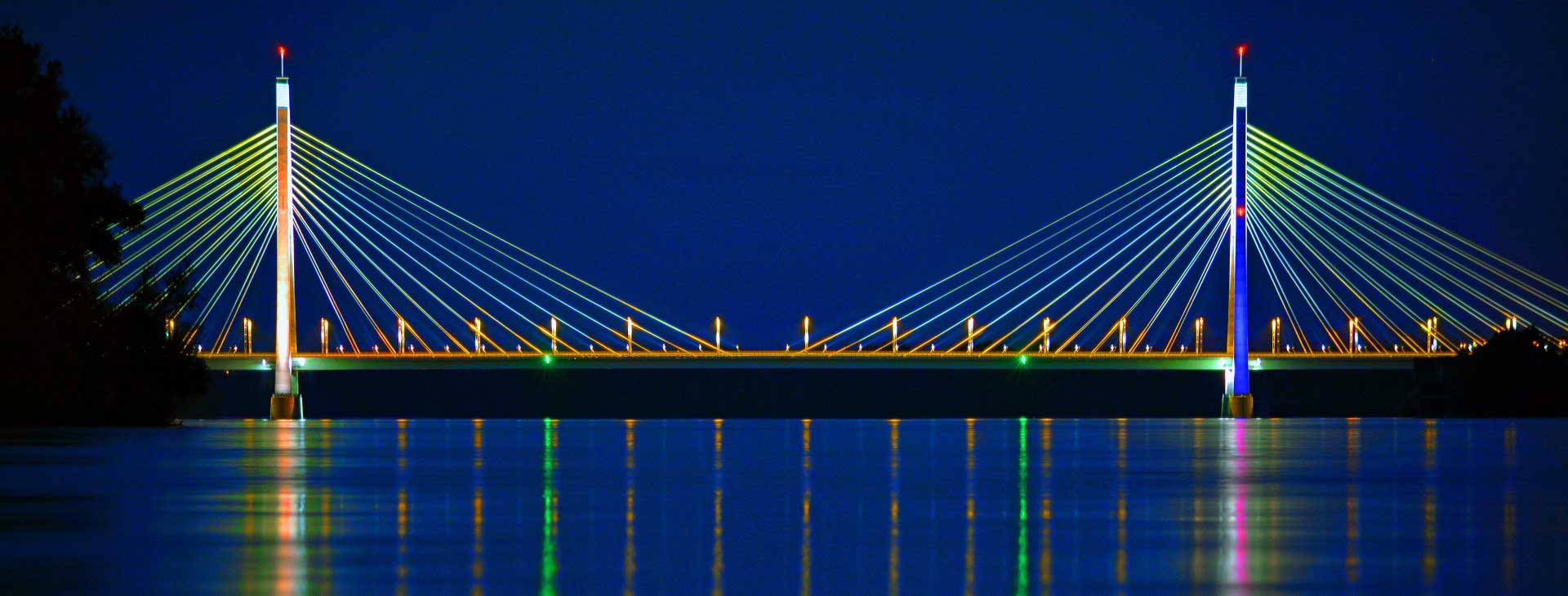 Eine Schrägseilbrücke, die nachts beleuchtet wird. Die Brücke hat zwei Türme mit Seilen, die sich zur Fahrbahn erstrecken. Die Brücke spiegelt sich im Wasser darunter.