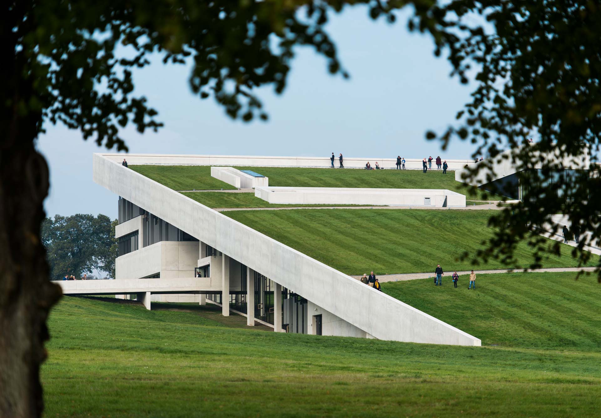 Ein modernes Gebäude mit einem einzigartigen architektonischen Design, das ein geneigtes, grasbewachsenes Dach aufweist, das bis zum Boden reicht und Zugang zum Dach ermöglicht. Dieses Design integriert die Struktur in die Landschaft.