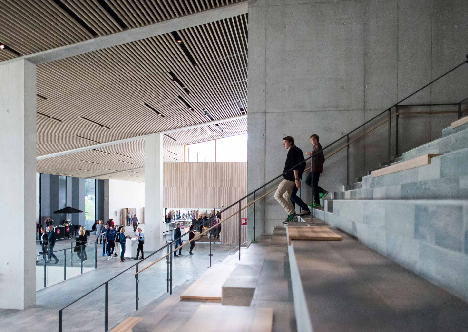 Ein Innenraum mit moderner Architektur zeigt eine Treppe mit Glasgeländern und Betonstufen. Personen steigen die Treppe hinauf, unter ihnen befindet sich ein geräumiger Bereich mit weiteren Personen, die an Tischen sitzen.