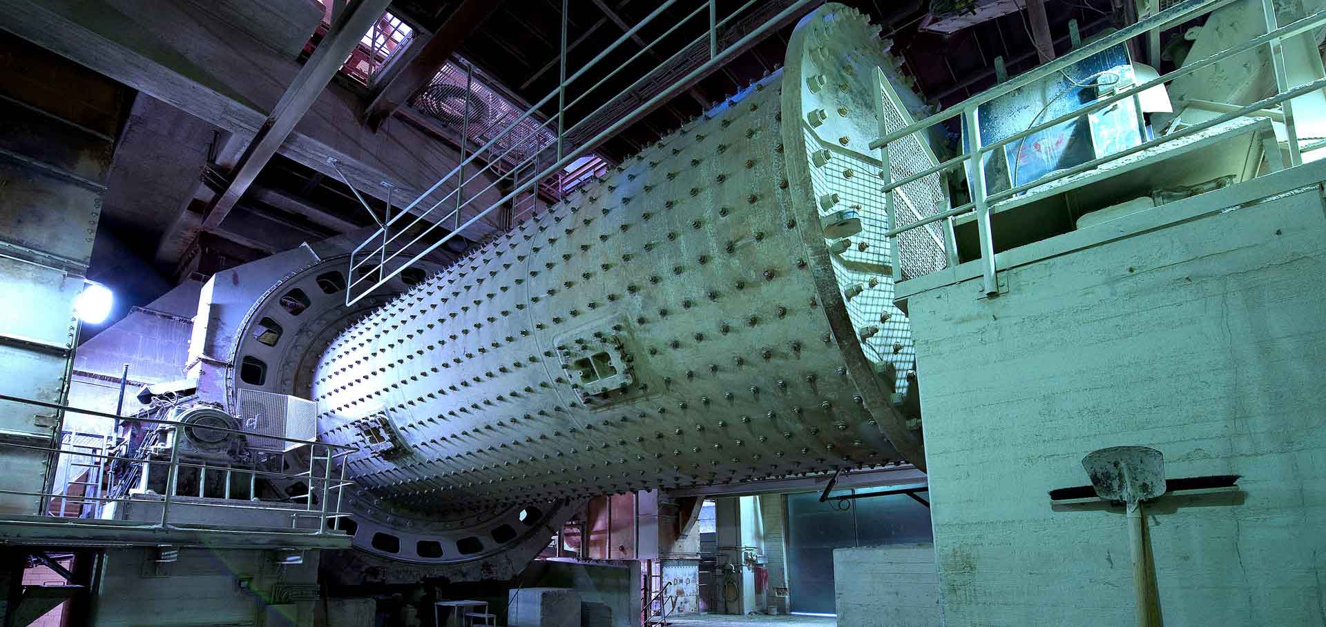 A large, industrial grinding mill with a cylindrical drum and a complex system of gears and pulleys. The mill is located in a large industrial hall with exposed beams and pipes.