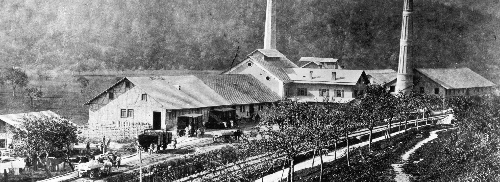 Historisches Foto industrieller Hallen mit Schloten, davor Güter-Waggons, Kutschen und Menschen