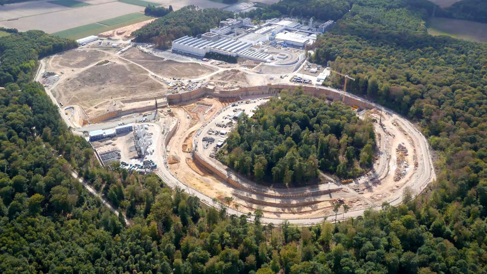 Aerial view of a large construction site with a circular excavation in the center, surrounded by buildings and heavy machinery.