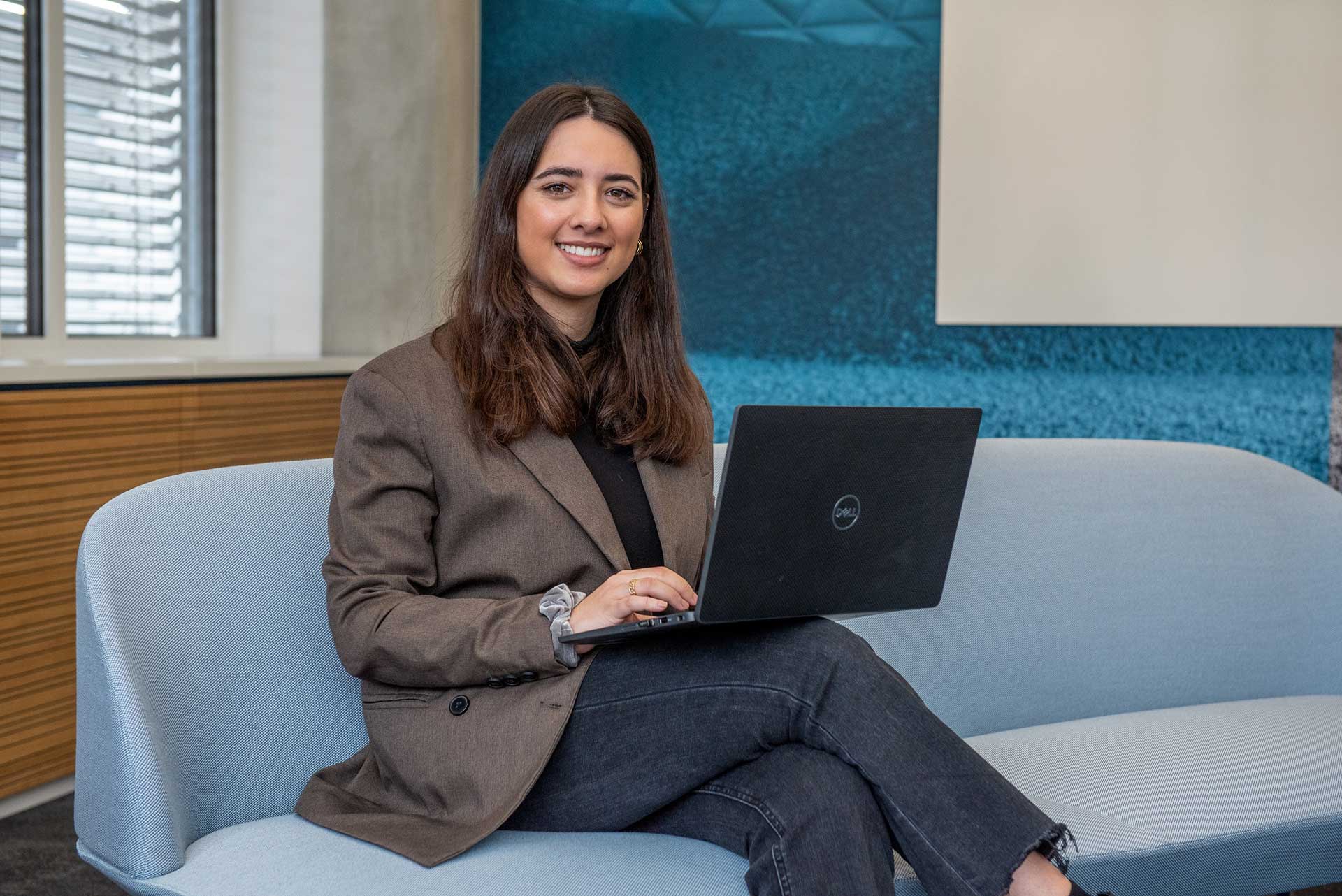 Junge Frau in Businesskleidung auf einem hellblauen Sofa, auf dem Schoß ein Laptop