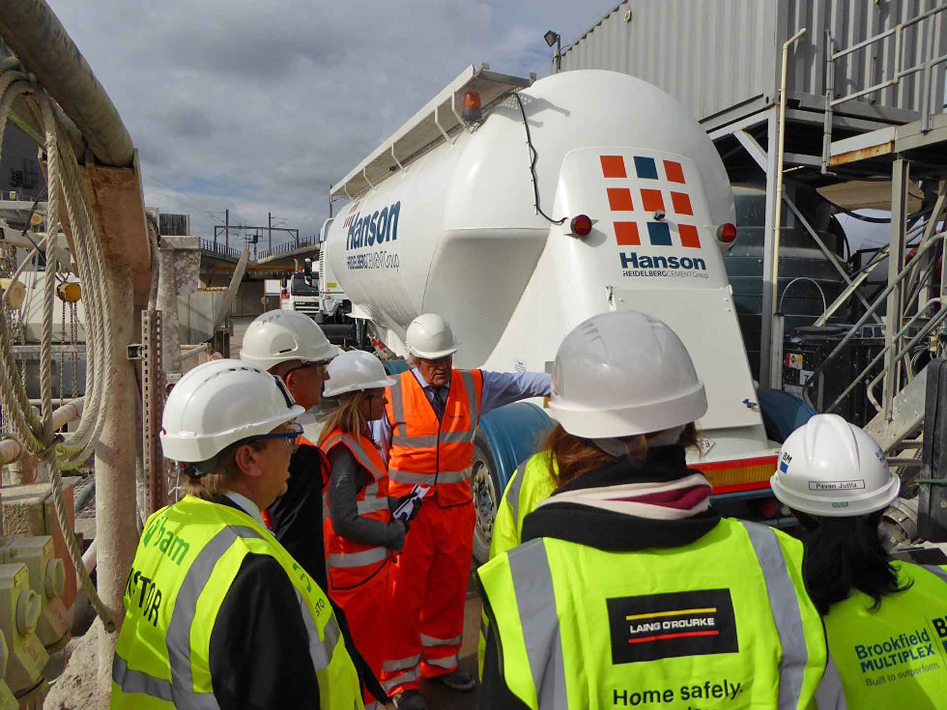 A group of people in safety gear attentively listening to another person by a ready-mixed concrete truck.