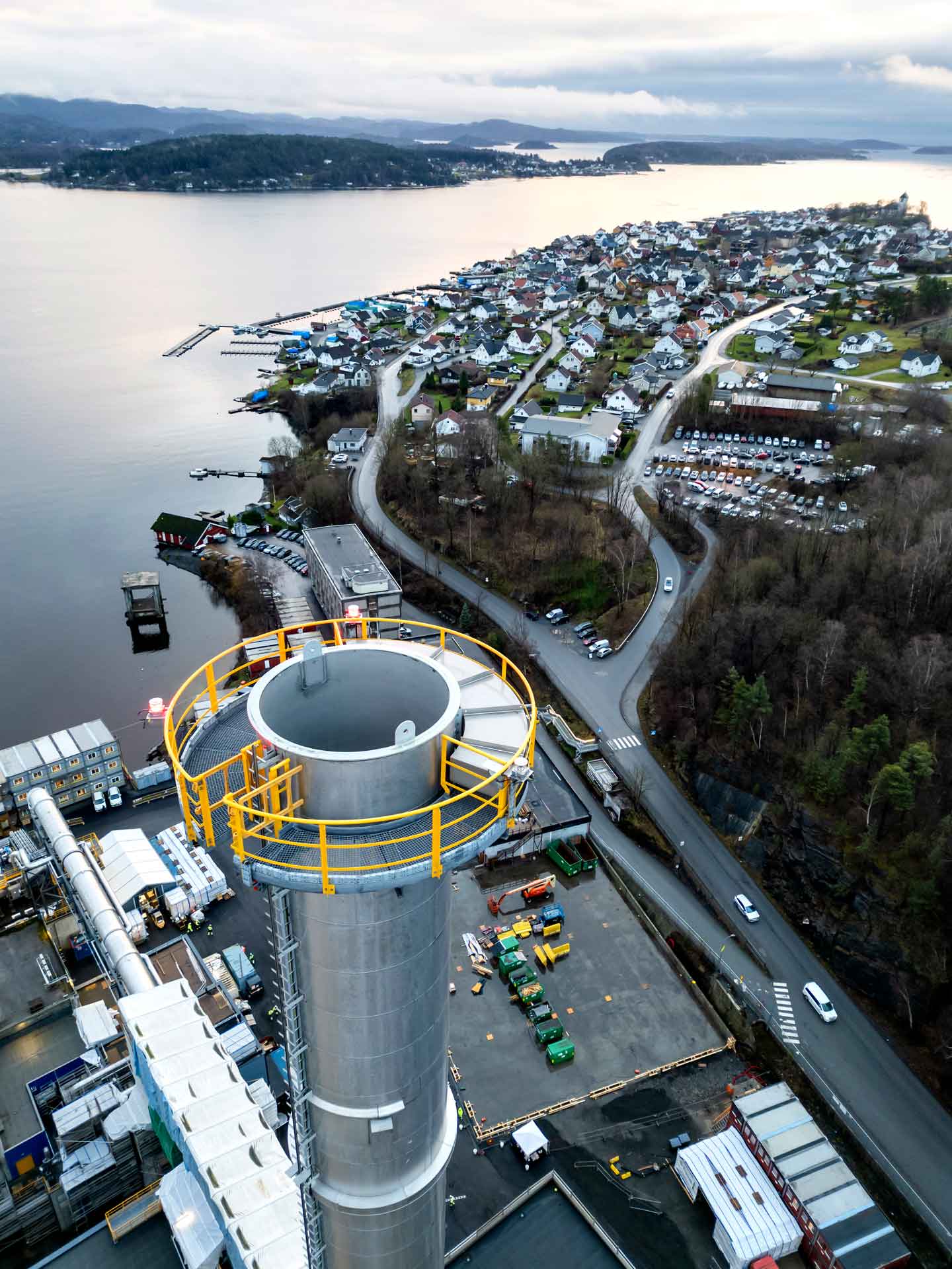 Luftaufnahme einer Küstenstadt mit einem großen Industrie-Schornstein im Vordergrund. Der Schornstein hat oben ein gelbes Sicherheitsgeländer.