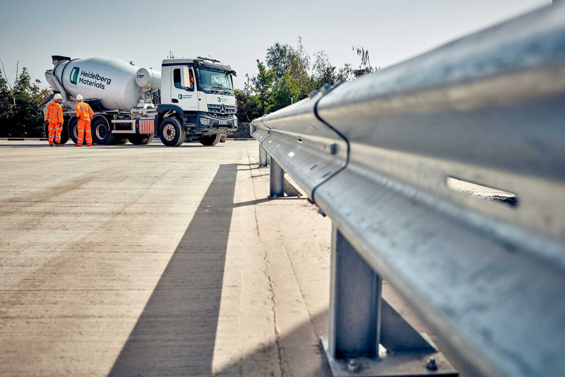 A ready-mixed concrete truck is parked on a paved surface, with two workers in safety vests standing nearby. A metal guardrail is visible in the foreground.