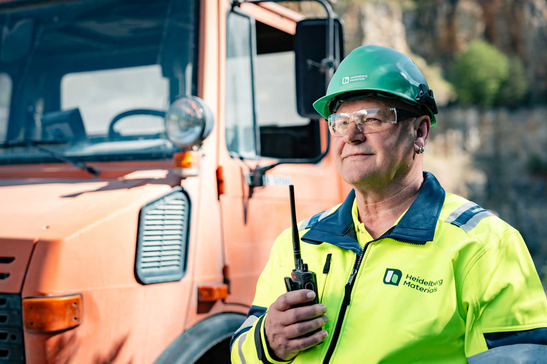 Man in yellow protective clothing and a green helmet stands next to an orange commercial vehicle and holds a walkie-talkie in his hand