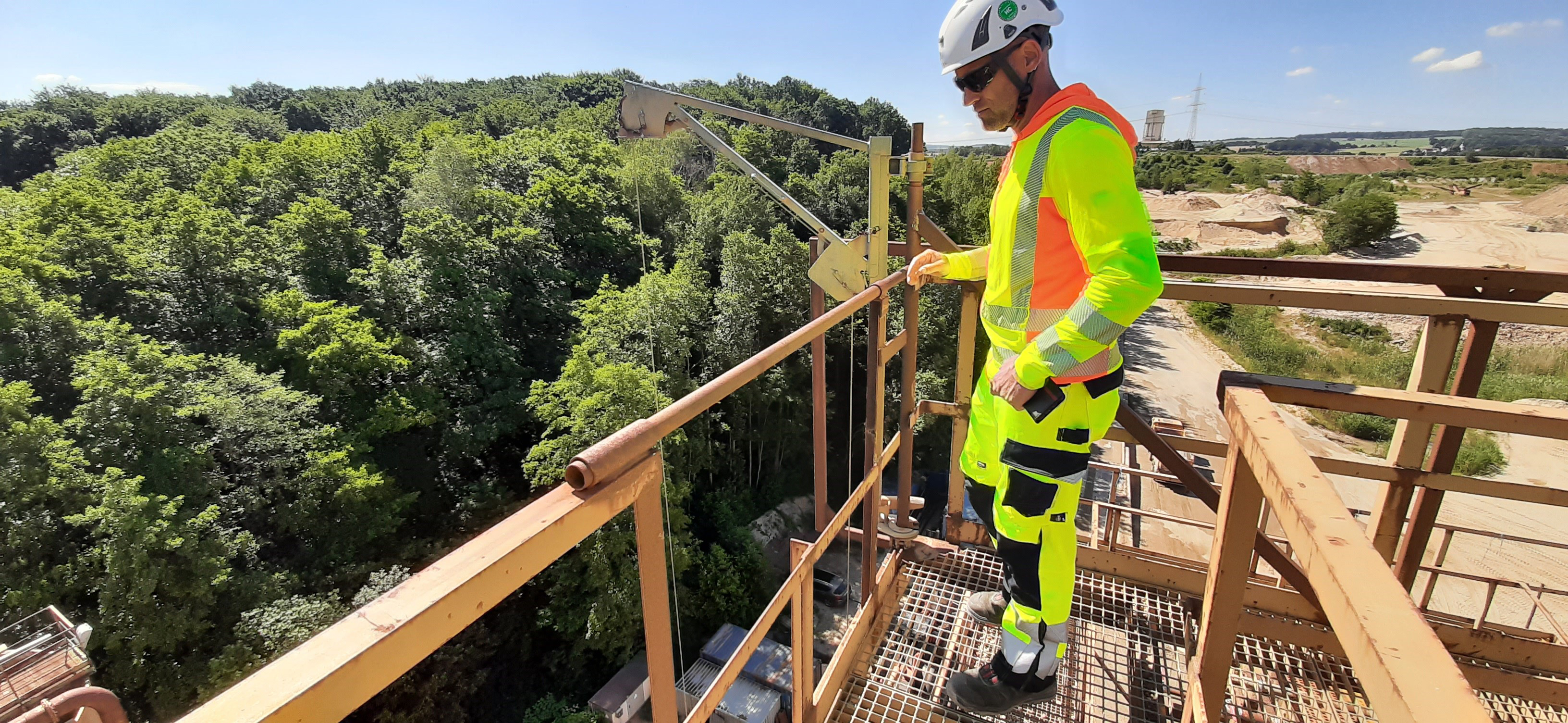 Man in protective clothing stands on a scaffolding, in the background forest and the area of a gravel plant
