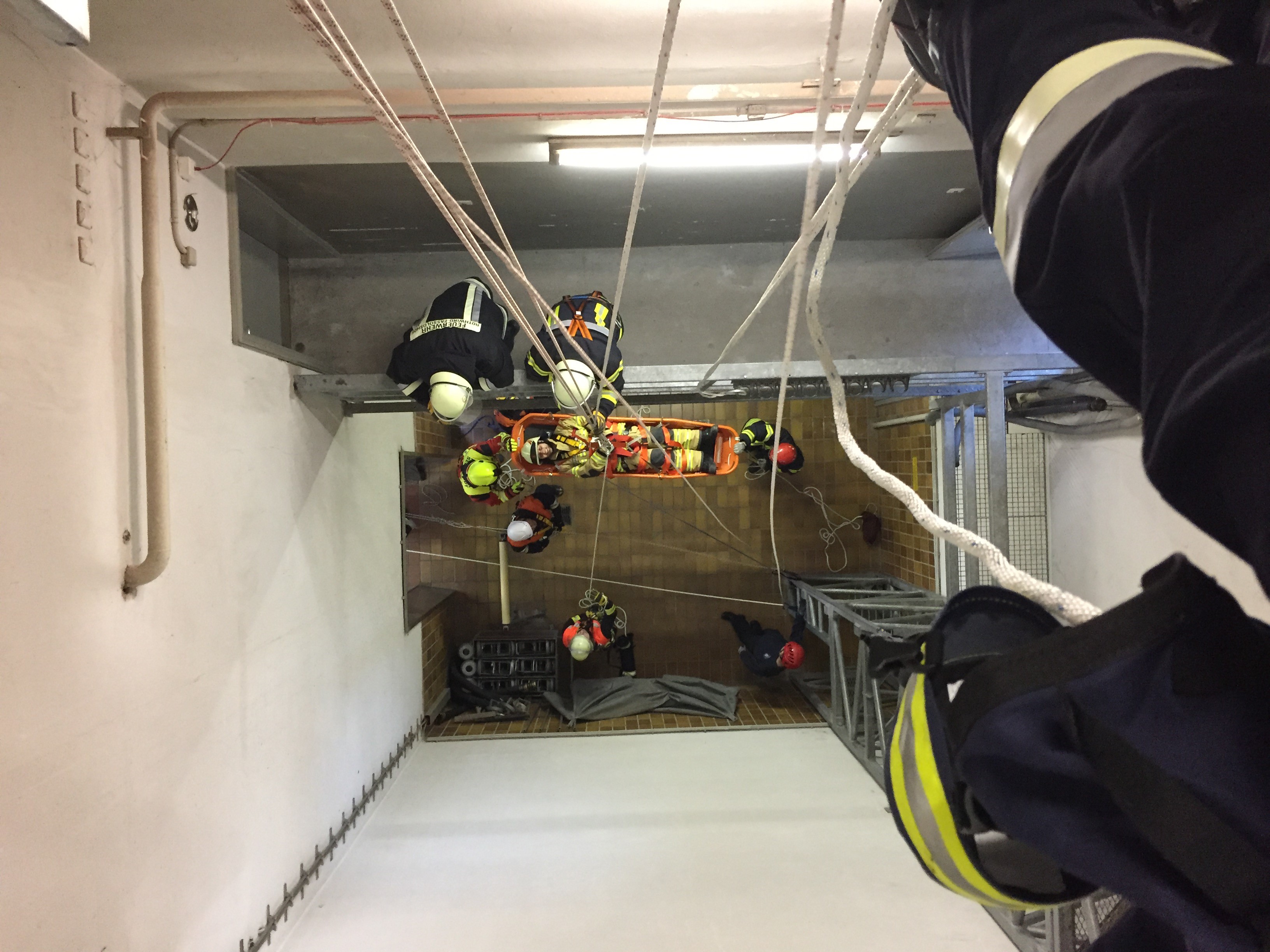 View from above into a shaft, on the floor of which a person has been placed in a stretcher, other people are busy lifting this stretcher upwards on ropes.