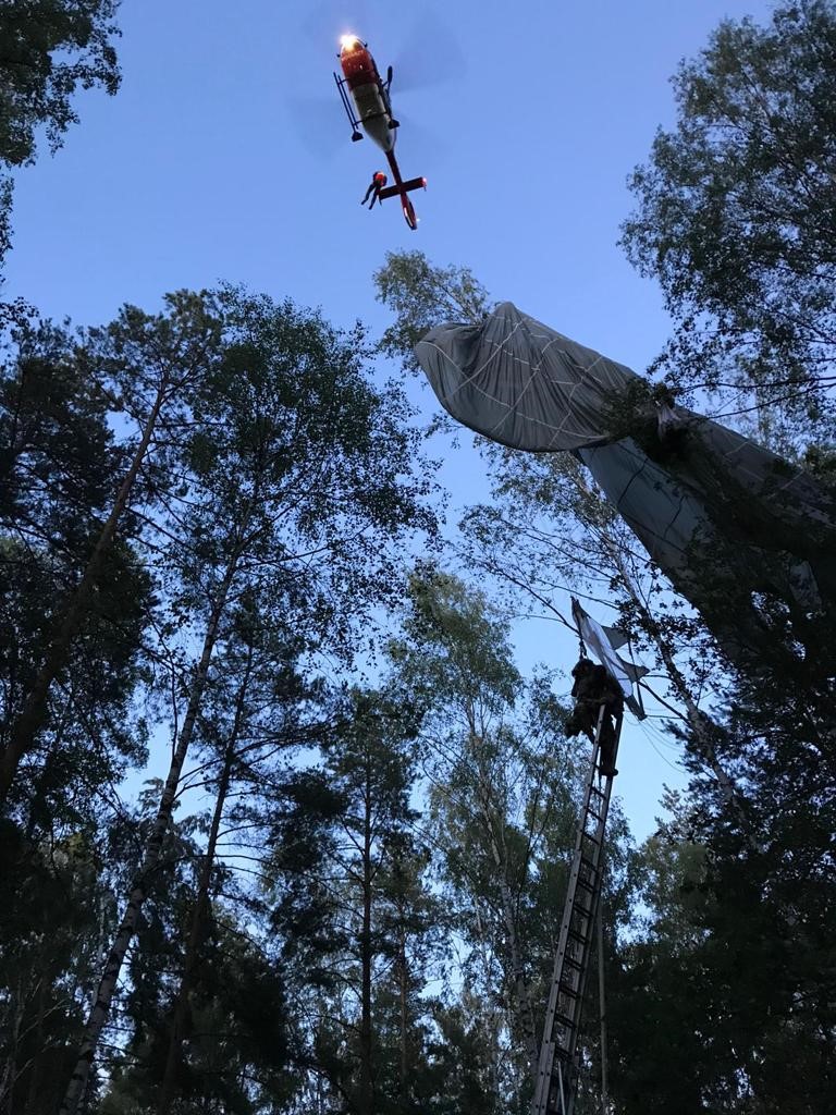 View from below between treetops into the sky. A parachute hangs from a treetop, with a helicopter above. A person climbs up a ladder to the parachute.