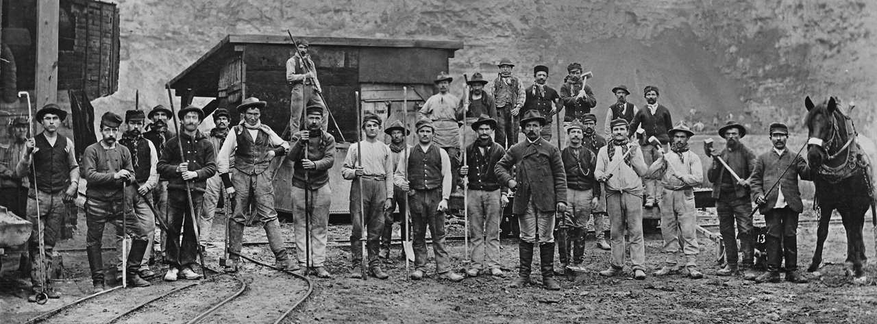 Historisches Foto; eine Gruppe von Männern mit Schaufeln und anderem Werkzeug steht vor einer Felswand in einem Steinbruch auf Schienen und schaut in die Kamera