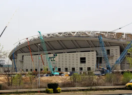 Weitläufige Baustelle eines großen Stadions, das die komplexe Struktur von Kränen und Gerüsten unter einem klaren Himmel präsentiert