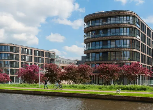Modern curved buildings alongside a canal with blooming pink trees, a clear blue sky, and people enjoying the outdoors