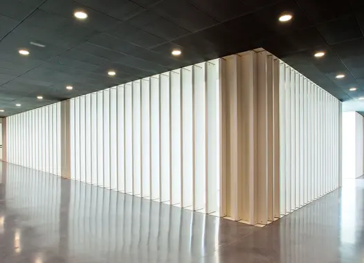 Ein modern gestaltetes Interieur mit einer großen, rechteckigen Struktur aus vertikalen weißen Paneelen, die Licht durchlassen und ein Muster auf dem glänzenden Boden erzeugen, unter einer dunklen Decke mit Einbauleuchten.