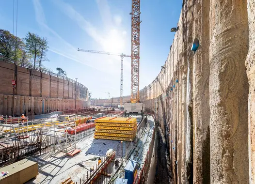 Construction site with deep excavation pit and retaining walls, featuring cranes and construction materials under a clear blue sky